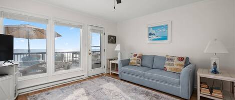 A living room with a sofa, a television, a ceiling fan, a large window with a view, and a rug.