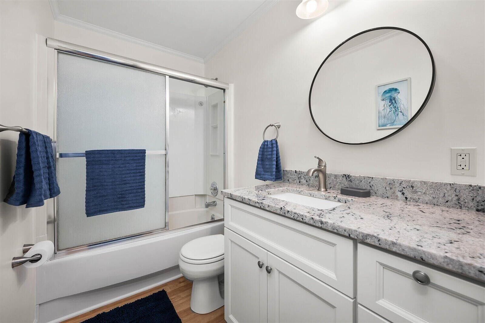 A modern bathroom with a glass shower door, a white toilet, a sink with a marble countertop, and a round mirror with a framed picture.