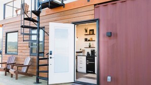 A modern cabin with a wooden exterior, a sliding glass door leading to a kitchen, and a small outdoor seating area with Adirondack chairs. - Somewhere in Texas Container Home (Alpine)