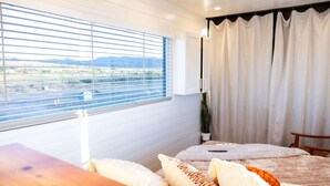 A bedroom with a large window, a bed with multiple pillows, a wooden nightstand, and a view of a landscape. - Somewhere in Texas Container Home (Alpine)
