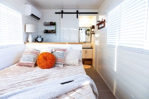 A bedroom with a bed, pillows, a bedside table, a lamp, a window, and a view of the bathroom.