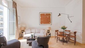 Un salon moderne comprenant un canapé, des fauteuils, une table basse, une table à manger avec des chaises, une lampe et une œuvre d’art encadrée accrochée au mur.