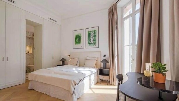 Une chambre à coucher avec un lit, un bureau et une chaise. Il y a une fenêtre avec des rideaux, une porte et un mur orné de deux tableaux encadrés.
