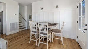 A dining area with a wooden table and four chairs, a white wall with paneling, a staircase in the background, and a door with a window.