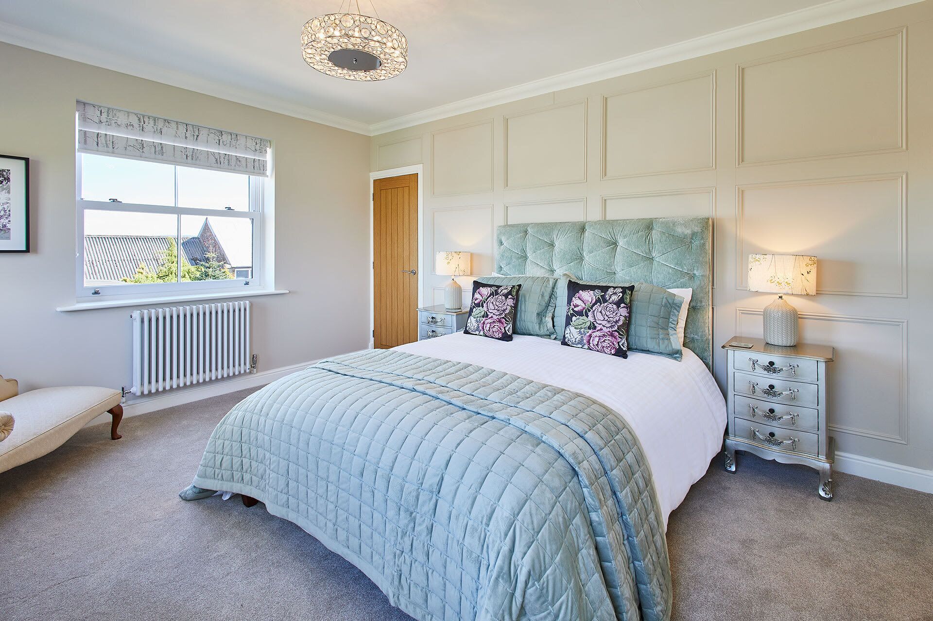 A bedroom with a large bed, a window with blinds, a radiator, a door, and a nightstand with a lamp.