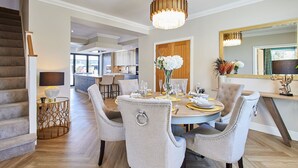 Un coin repas avec une table ronde dressée pour six personnes, entourée de chaises rembourrées ornées de motifs décoratifs. Un lustre est suspendu au-dessus de la table, et un miroir à cadre doré est accroché au mur, à droite.