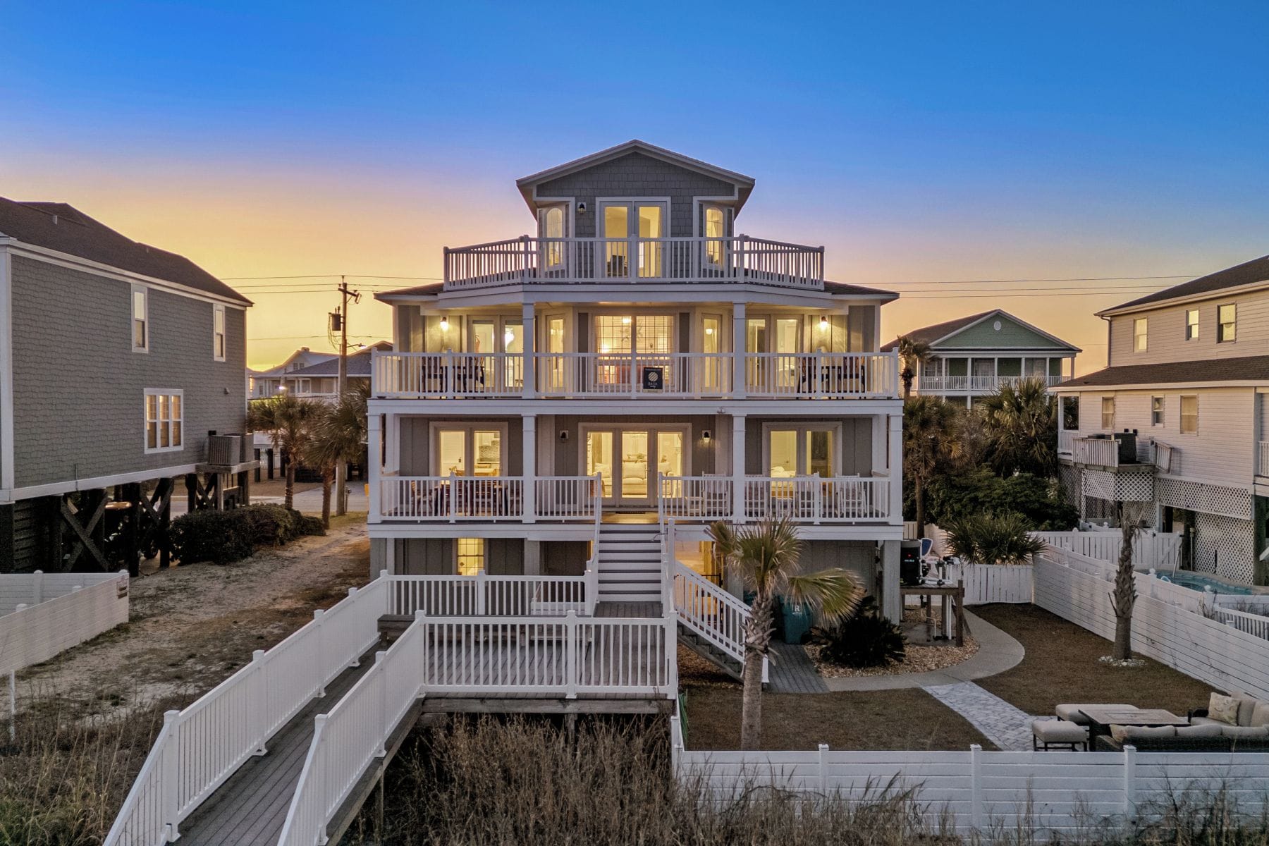 Une maison à plusieurs étages située en bord de mer, dotée d’un balcon à chaque étage, d’un porche couvert et d’un petit espace extérieur aménagé pour s’asseoir.