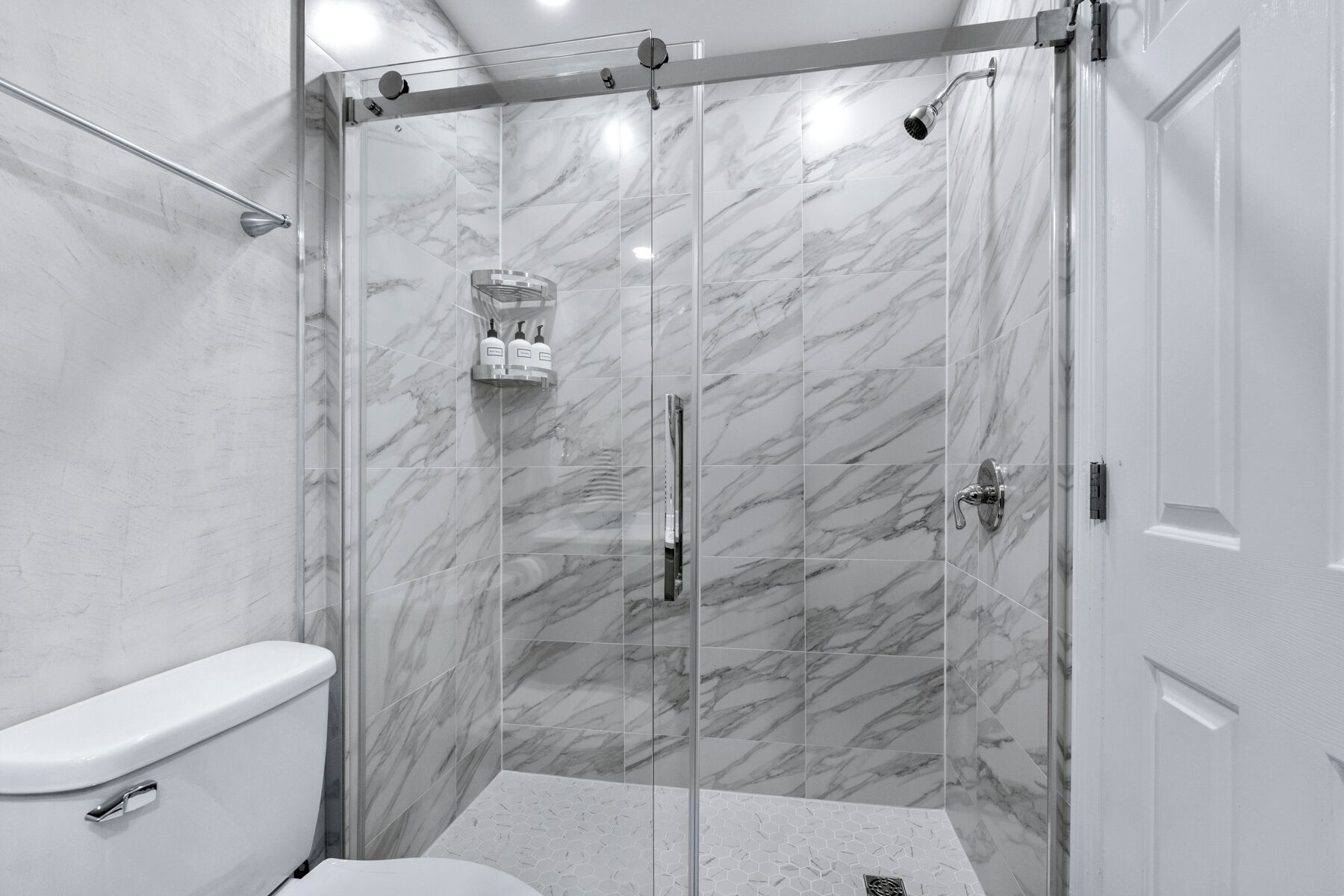Un baño moderno con ducha envuelta en vidrio, azulejos de mármol, inodoro y lavamanos con dispensadores de jabón.