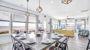 A modern dining room with a large table set for six, large windows with blinds, and a view of the beach.