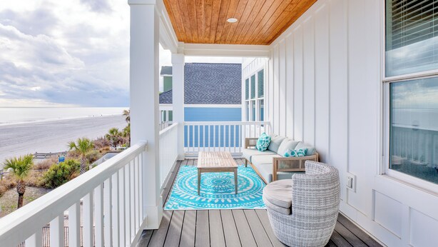 A balcony with a wooden ceiling, white railings, a blue patterned rug, a wicker chair, and a sofa with cushions.
