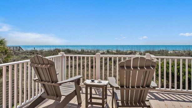 Eine Holzterrasse mit zwei Adirondack-Stühlen und einem kleinen Tisch mit Blick auf einen Strand und klares, blaues Wasser.