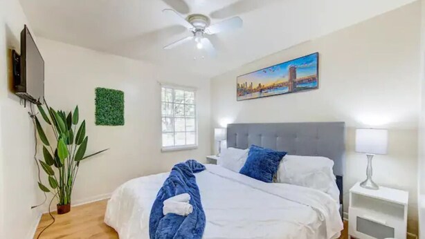 Una habitación de hotel con una cama, ventilador de techo, un cuadro, ventana y una planta.