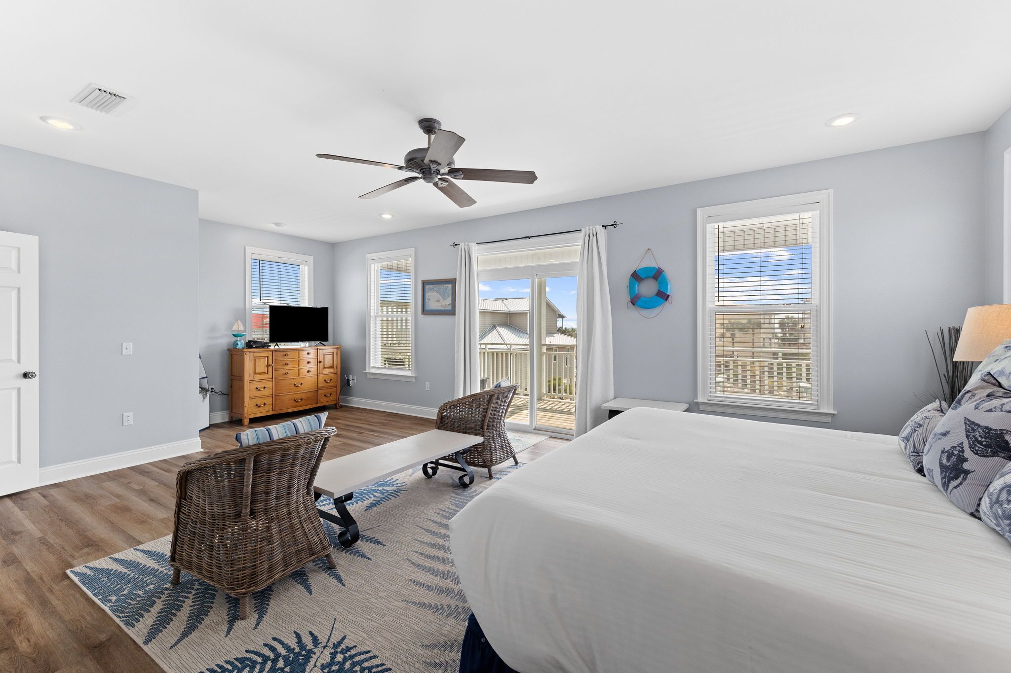 Une chambre avec un lit, un ventilateur de plafond, une commode, un fauteuil en osier, une petite table et une vue sur l’extérieur.