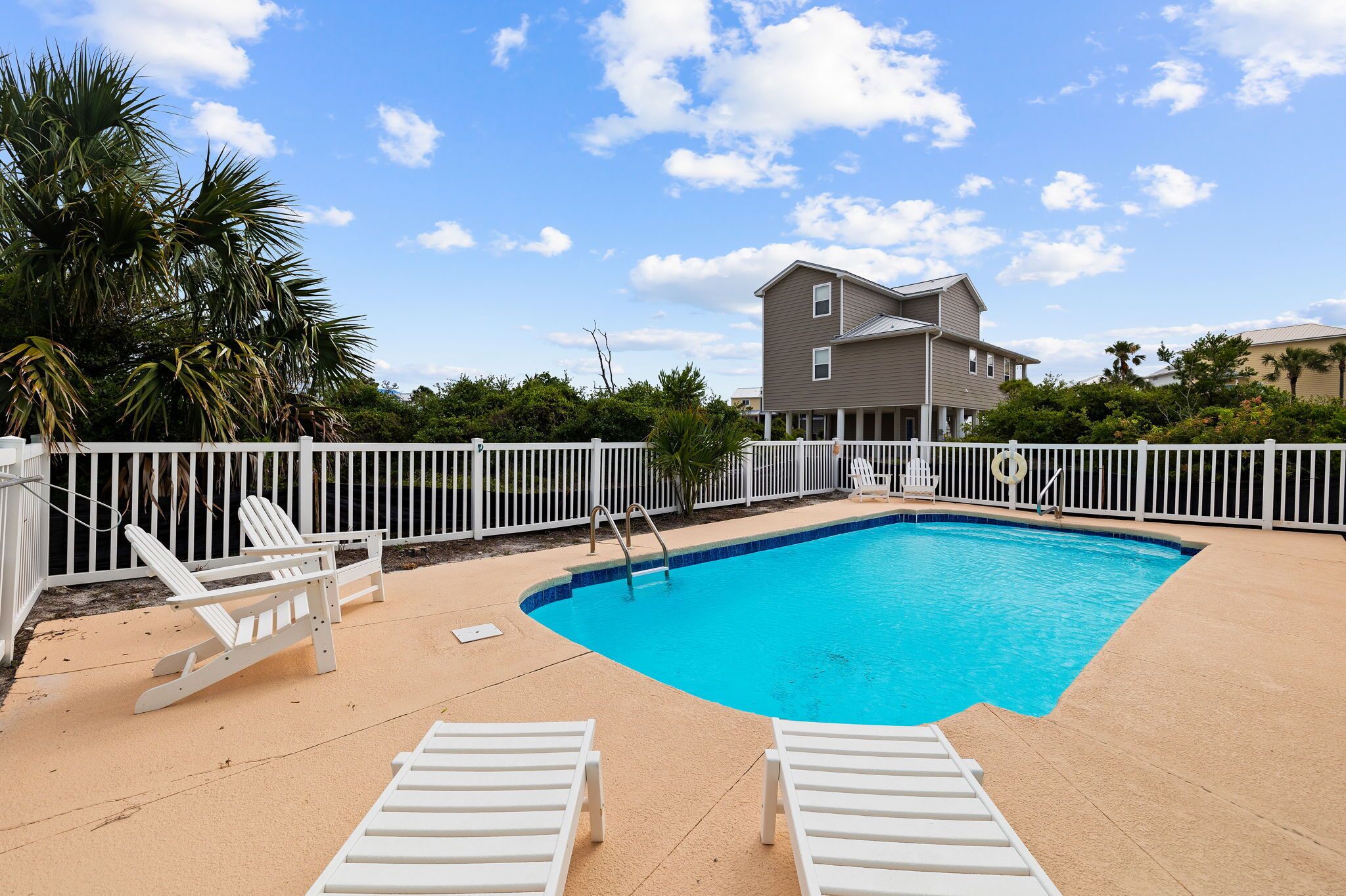 A swimming pool with white lounge chairs, a white fence, and a two-story building in the background.