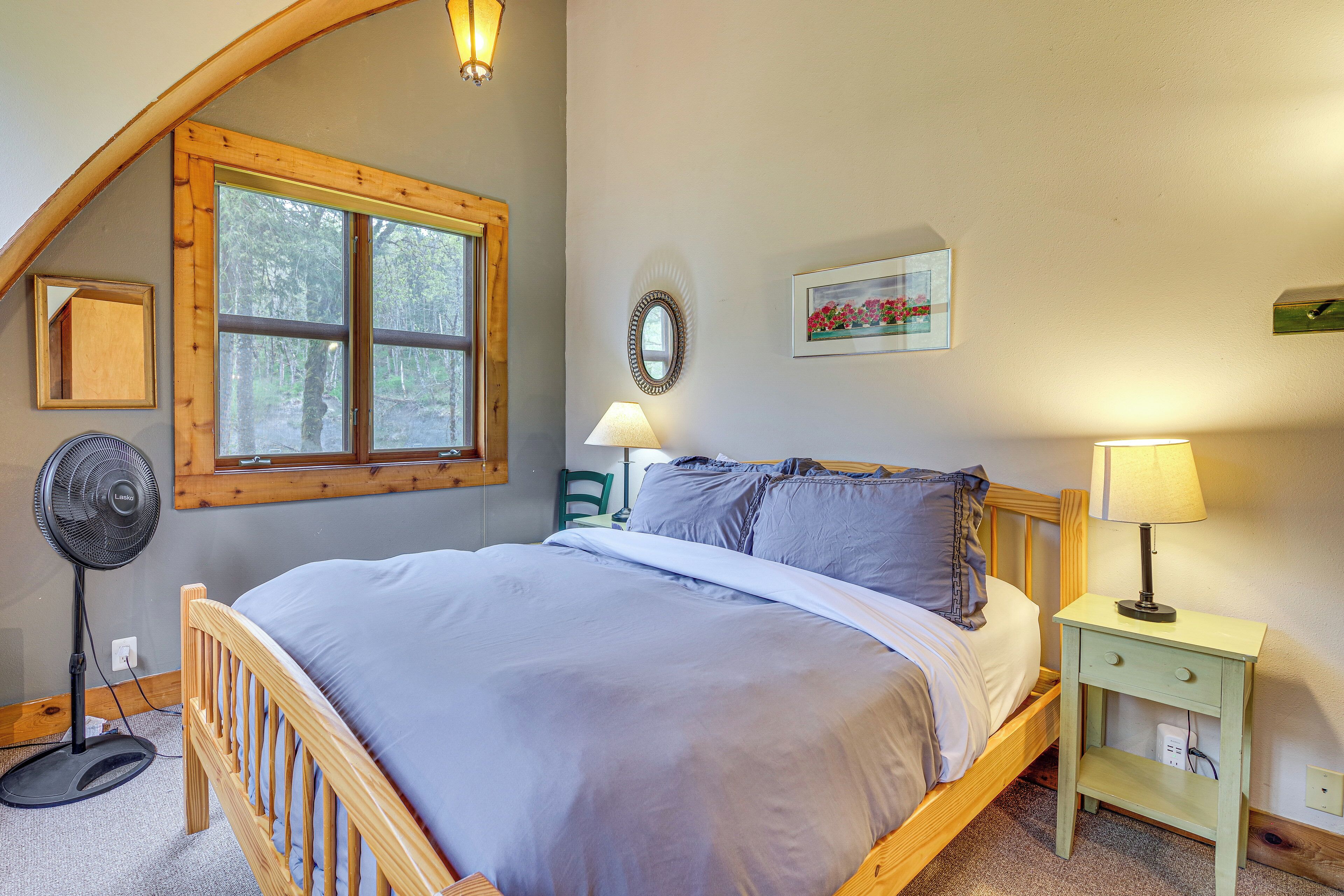 A bedroom with a wooden bed, a standing fan, a nightstand with a lamp, a window with a view of trees, and a mirror on the wall.