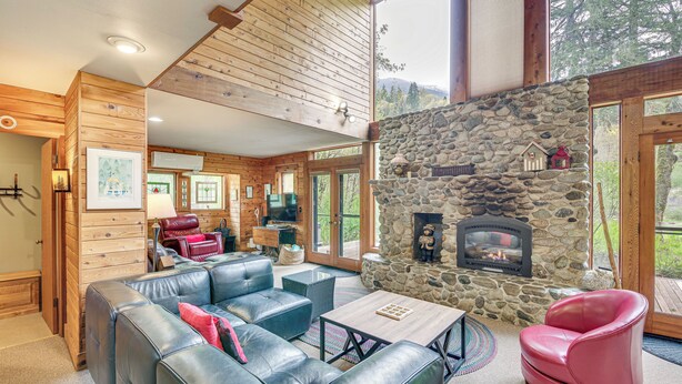 A log cabin interior with a stone fireplace, leather furniture, and a large window overlooking a forested area. - Riverfront Skykomish Cabin w/ Private Hot Tub! (Skykomish)