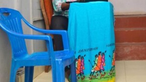 A room with a wooden door, a blue plastic chair, and a table with a colorful cloth. - Rafiki Apartment Hotel (Zanzibar Town)