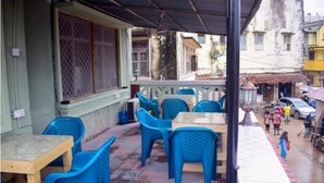 An outdoor seating area with blue plastic chairs and tables, a covered section with a metal roof, and a view of the street with pedestrians and buildings. - Rafiki Apartment Hotel (Zanzibar Town)