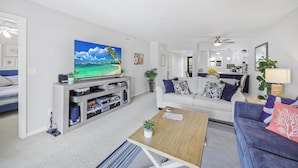 A modern living room with a flat-screen TV, a sofa, a coffee table, and a kitchen area in the background. - Rare Walkout Condo | Pool & Steps to Beach/Food (Marco Island)