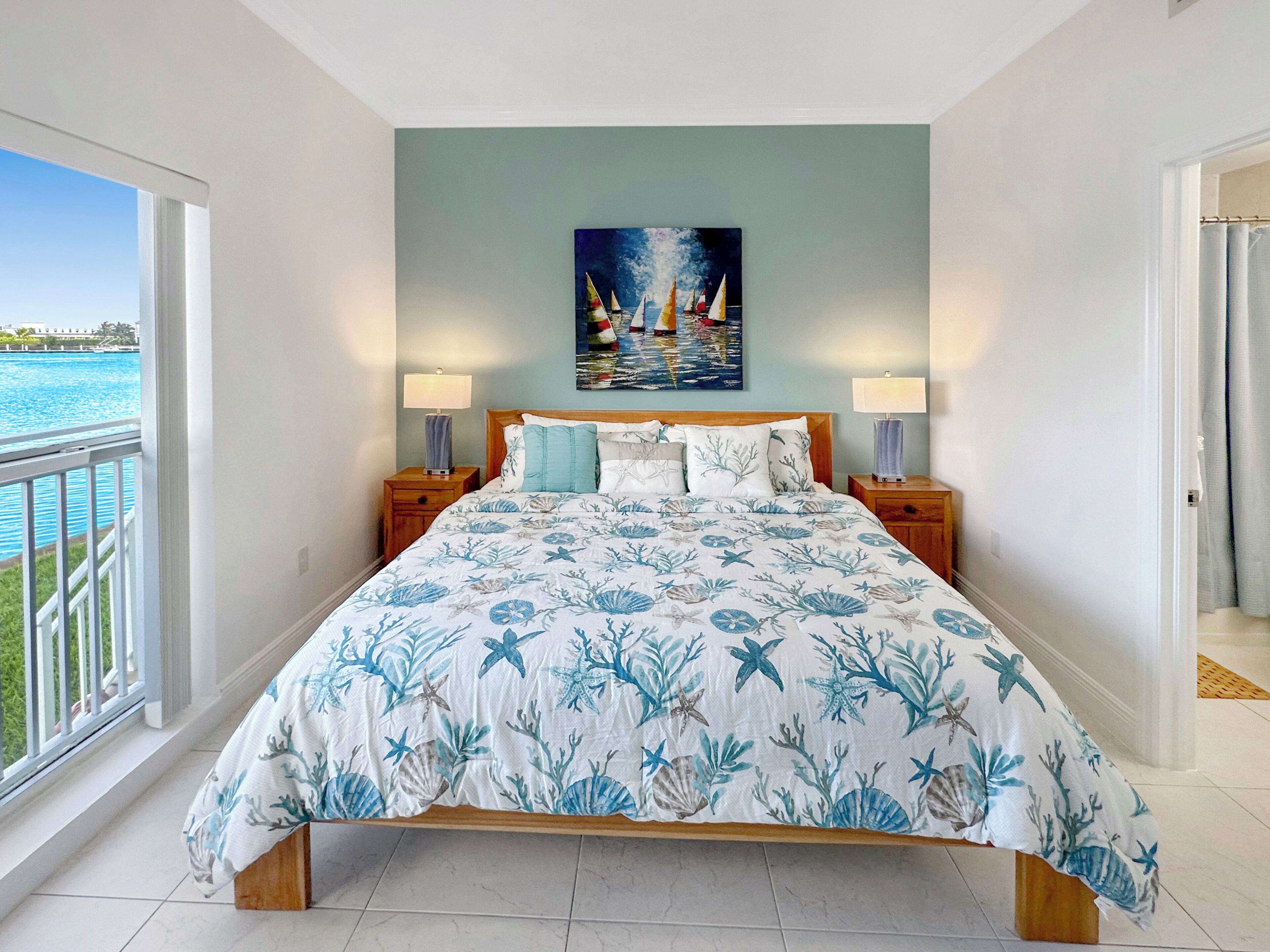 A bedroom with a bed, two bedside tables, a painting, and a view of the water.
