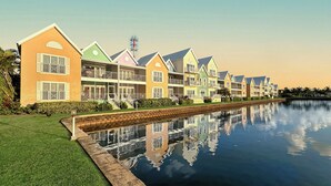 A row of colorful, multi-story buildings with balconies, a calm water body in the foreground, and a clear sky. - Cayman Blu 2 Bedroom Waterfront Condo (Grand Harbour)