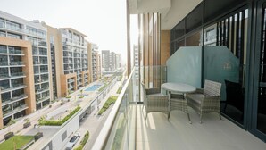 Un balcon doté de meubles en osier et d’une rambarde en verre, offrant une vue sur un quartier résidentiel avec des immeubles modernes et une piscine.