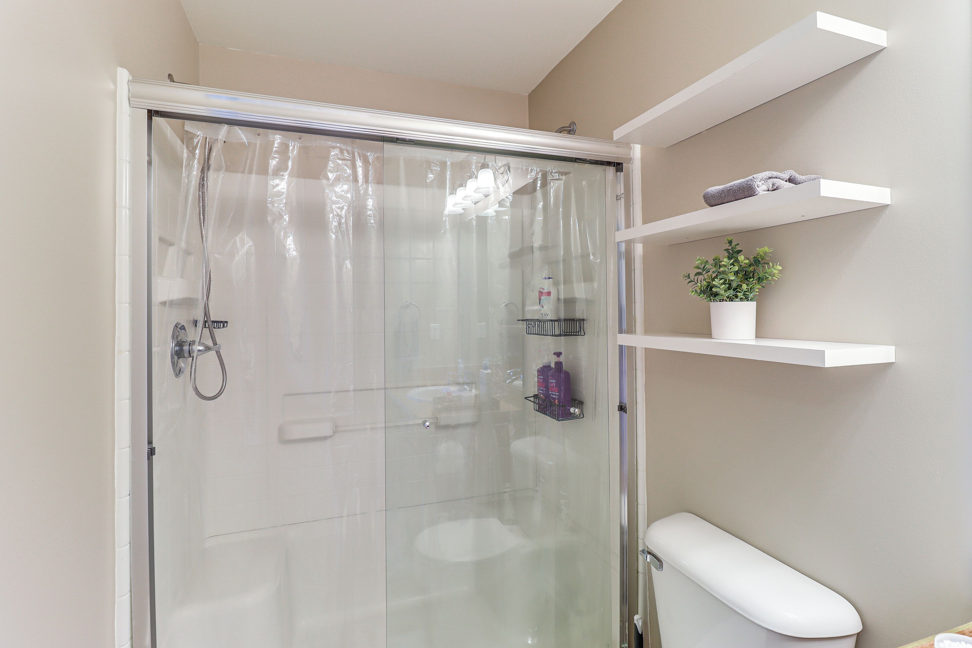 Un baño moderno con cabina de ducha de vidrio, inodoro y estantes blancos con artículos de aseo y una planta en maceta.