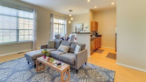Ein Wohnzimmer mit grauem Sofa, Couchtisch und einem gemusterten Teppich. Im Hintergrund ist eine Küchenzeile mit Holzschränken und einer Arbeitsfläche zu sehen.