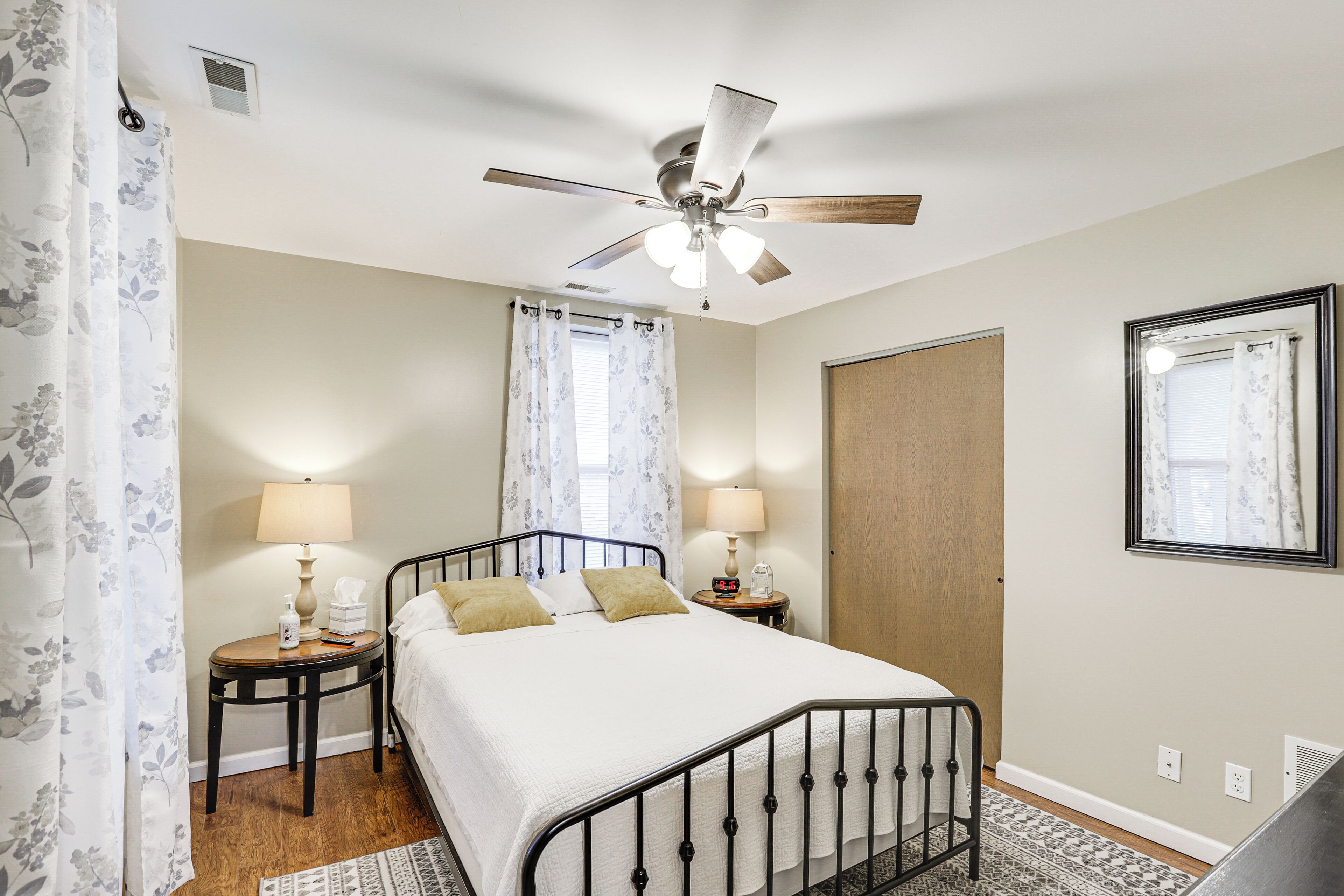 A bedroom with a bed, two bedside tables with lamps, a ceiling fan, a window with curtains, and a door.