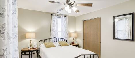 A bedroom with a bed, two bedside tables with lamps, a ceiling fan, a window with curtains, and a door.