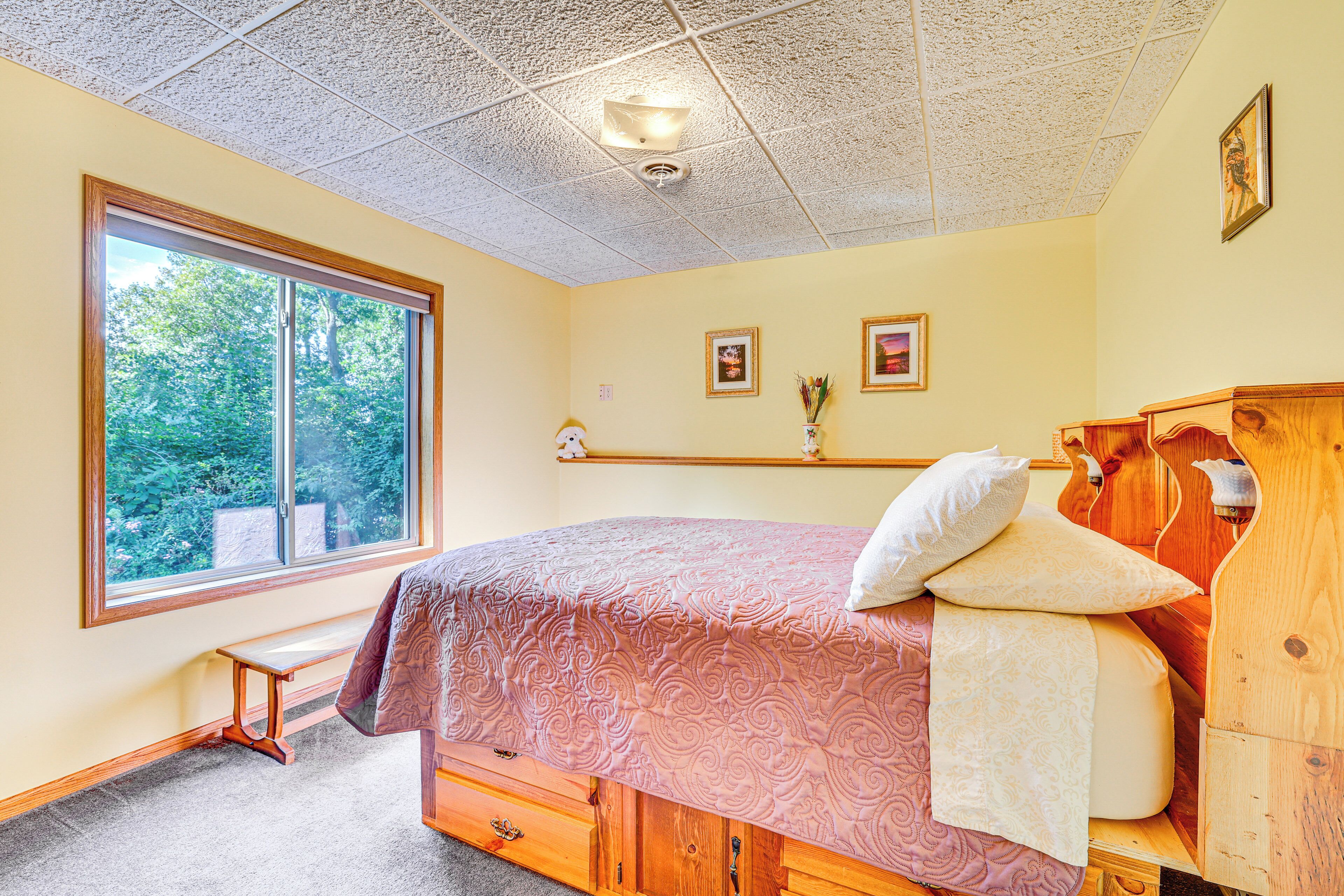 Une chambre avec une grande fenêtre, un lit en bois, un banc et une commode en bois.