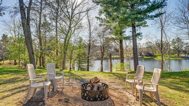 Un terrain de camping avec un foyer entouré de chaises, un lac et des arbres en arrière-plan.