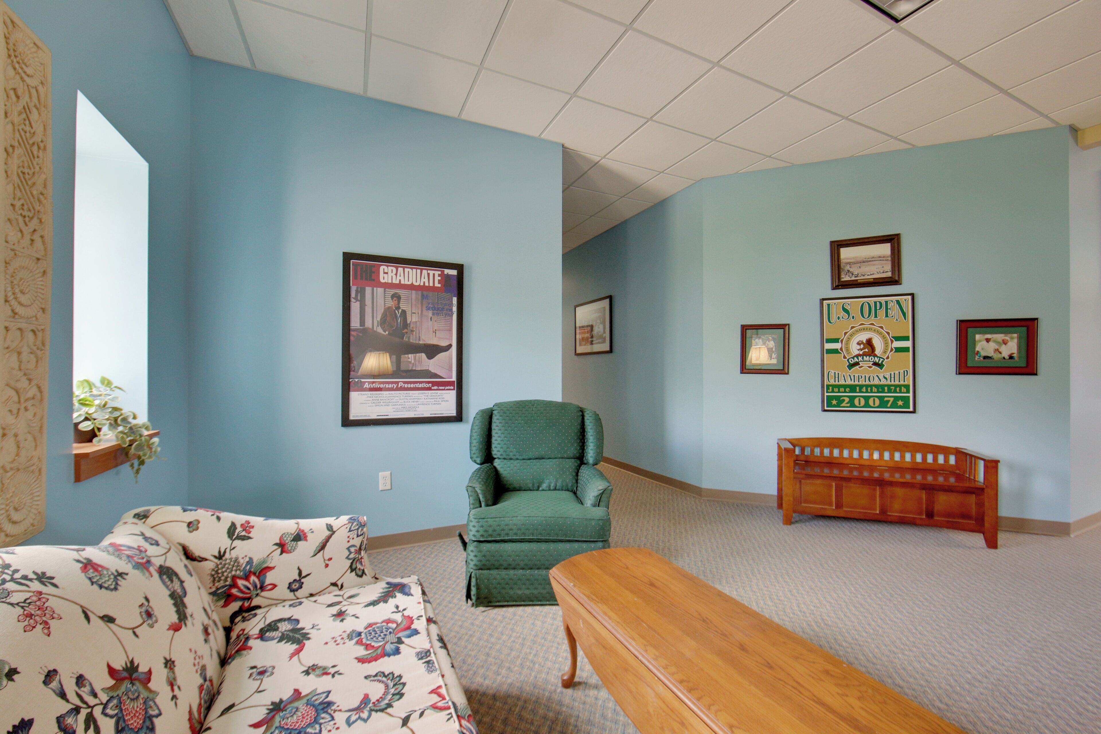 A room with a floral-patterned sofa, a green armchair, a wooden bench, and a wall with framed pictures and a poster.