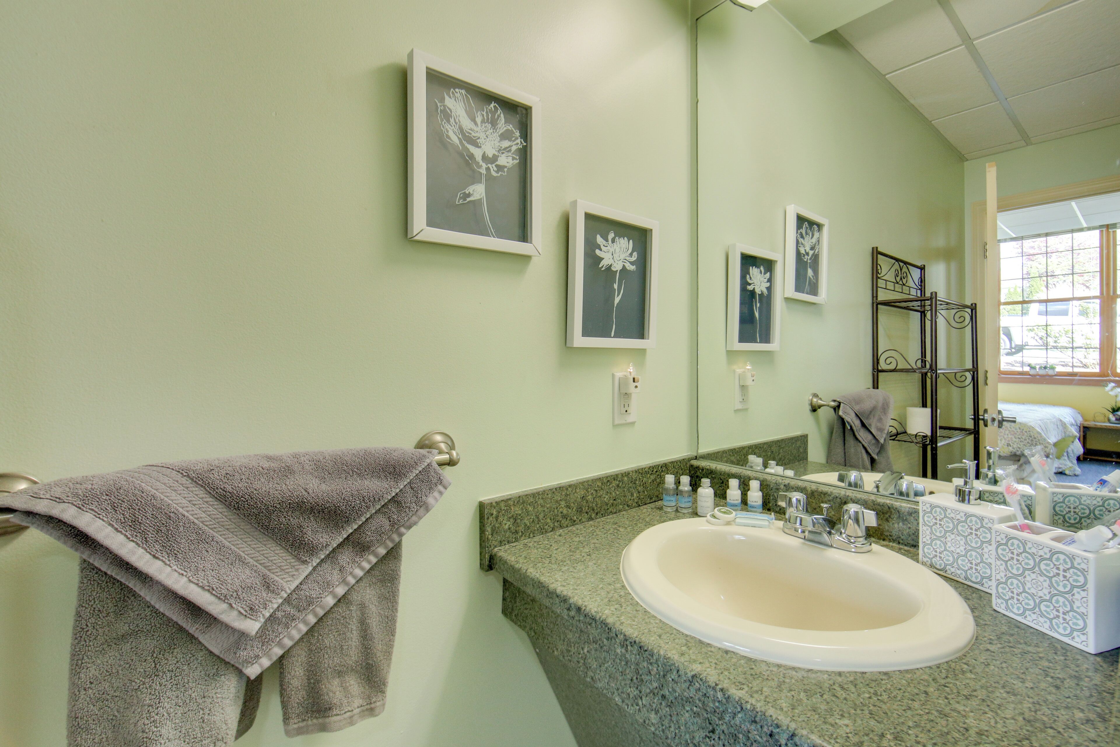 Une salle de bain avec un comptoir en granit, un lavabo, un miroir et des œuvres d’art florales encadrées accrochées au mur.