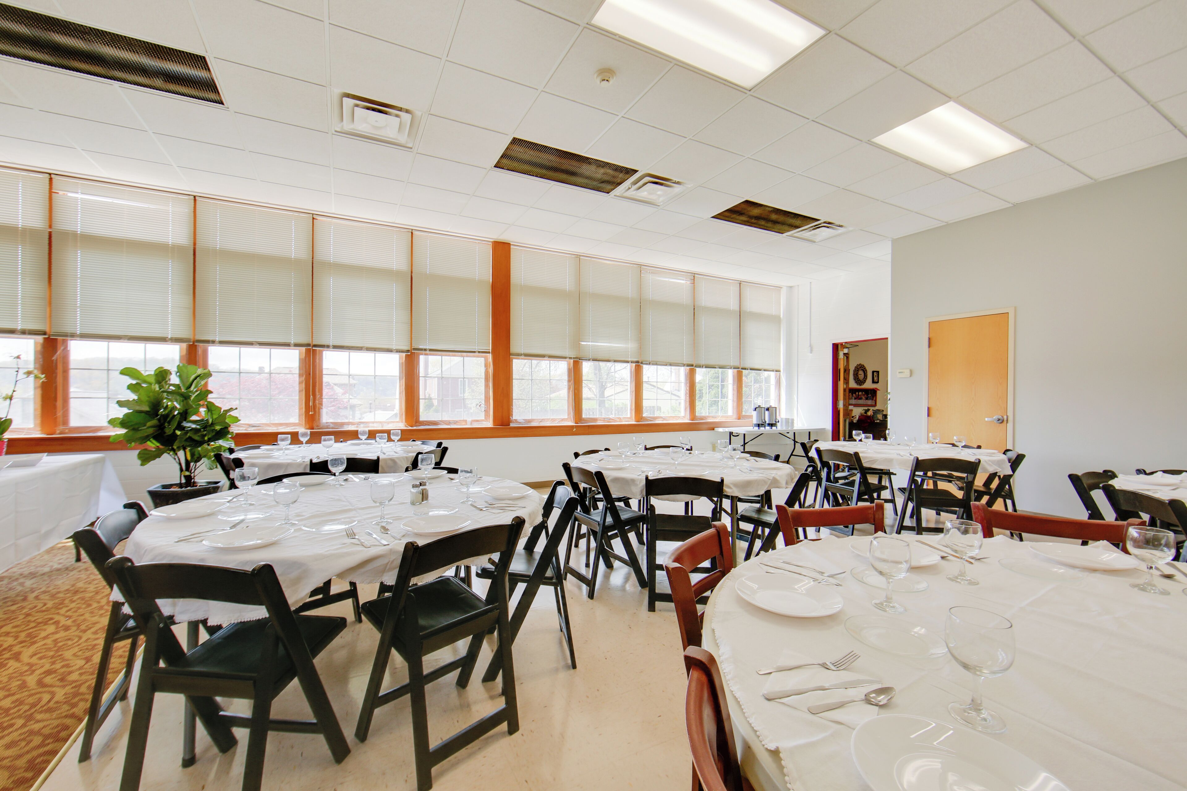 Un espace repas avec plusieurs tables dressées pour les repas, de grandes fenêtres équipées de stores et une plante dans un coin.