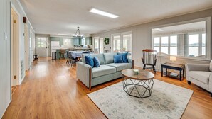 Ein geräumiges Wohnzimmer mit Holzdielen, einem grauen Sofa mit blauen Kissen, einem runden Couchtisch und Blick zur Küche im Hintergrund.