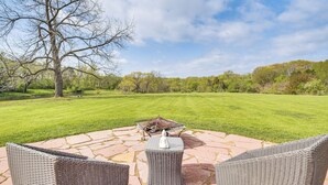 A patio with wicker furniture and a fire pit, overlooking a well-manicured lawn and trees.