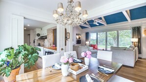 A spacious living area with a wooden dining table set for four, a chandelier, and a view of the outdoors through large windows.