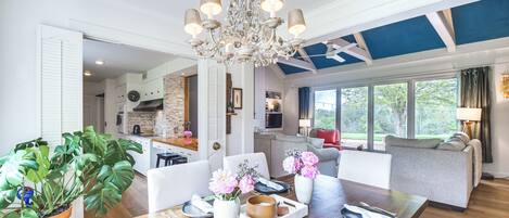 A spacious living area with a wooden dining table set for four, a chandelier, and a view of the outdoors through large windows.