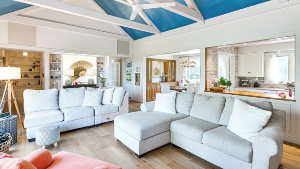 A spacious living area with a large sectional sofa, a small ottoman, and a pink chair. There is a ceiling fan, a large mirror, and a kitchen area visible in the background.