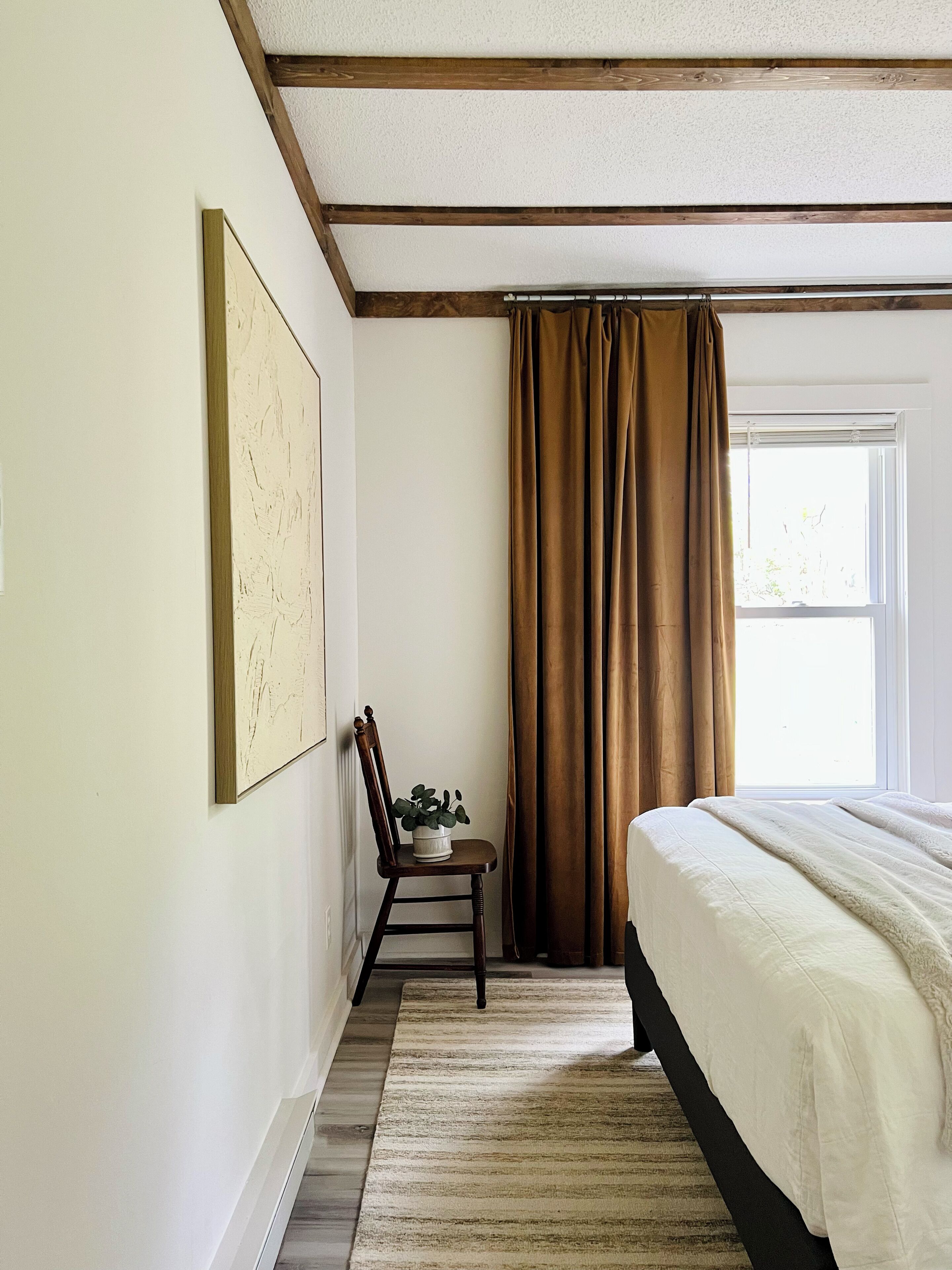 Une chambre à coucher avec un lit, une chaise agrémentée d’une plante, une fenêtre avec des rideaux et un tableau accroché au mur.