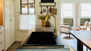 Une cuisine moderne avec un îlot central, un évier noir et un robinet doré. On y trouve une fenêtre avec des rideaux, une étagère avec des objets décoratifs et un coin repas avec une table et des chaises.