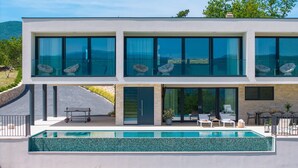 Een modern huis met twee verdiepingen, een groot raam op de bovenverdieping en een zwembad op de begane grond.