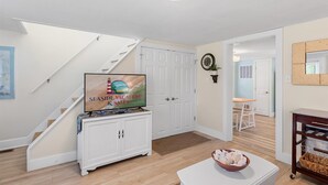 A spacious living room with a staircase, a white cabinet, a television displaying 'Seaside Vacations & Sales', and a dining area with a table and chairs. - Adorable Chincoteague home by Seaside Vacations & Sales (Chincoteague Island)