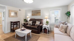 A living room with a brown leather sofa, a white coffee table, a beige sofa, and a white side table with a lamp. There is a painting on the wall and a window with blinds. - Adorable Chincoteague home by Seaside Vacations & Sales (Chincoteague Island)