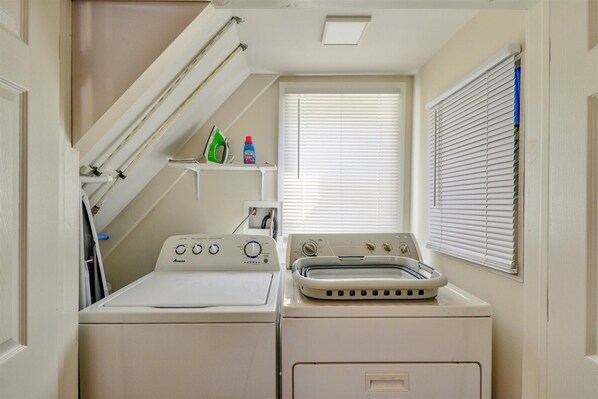 A compact laundry room with a front-loading washer and dryer, a drying rack, and a window with blinds. - Adorable Chincoteague home by Seaside Vacations & Sales (Chincoteague Island)