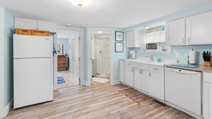A modern kitchen with white cabinets, a refrigerator, and a dishwasher. There is a wooden floor and a colorful rug. - Adorable Chincoteague home by Seaside Vacations & Sales (Chincoteague Island)