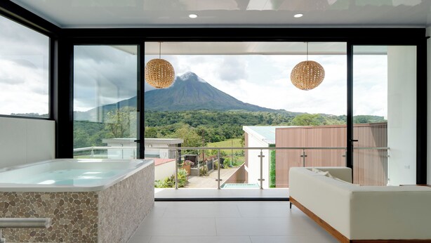 Ein modernes Badezimmer mit einer Eckbadewanne aus Steinmosaik, einer weißen Couch und einer großen Glasdoor, die auf einen Balkon mit Bergblick führt.