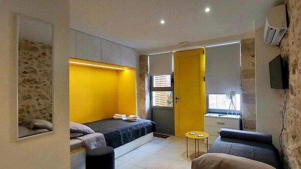 Une chambre d’hôtel de taille réduite, comprenant un lit, une petite table, un canapé, un miroir et une fenêtre avec des stores.