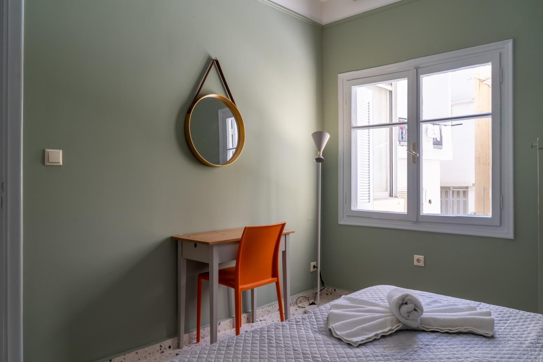 A bedroom with a bed, a desk, an orange chair, a window, and a wall-mounted mirror.
